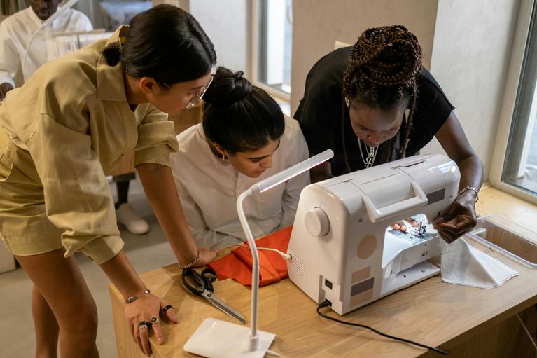 Three students surround a sewing machine to learn how to sew.